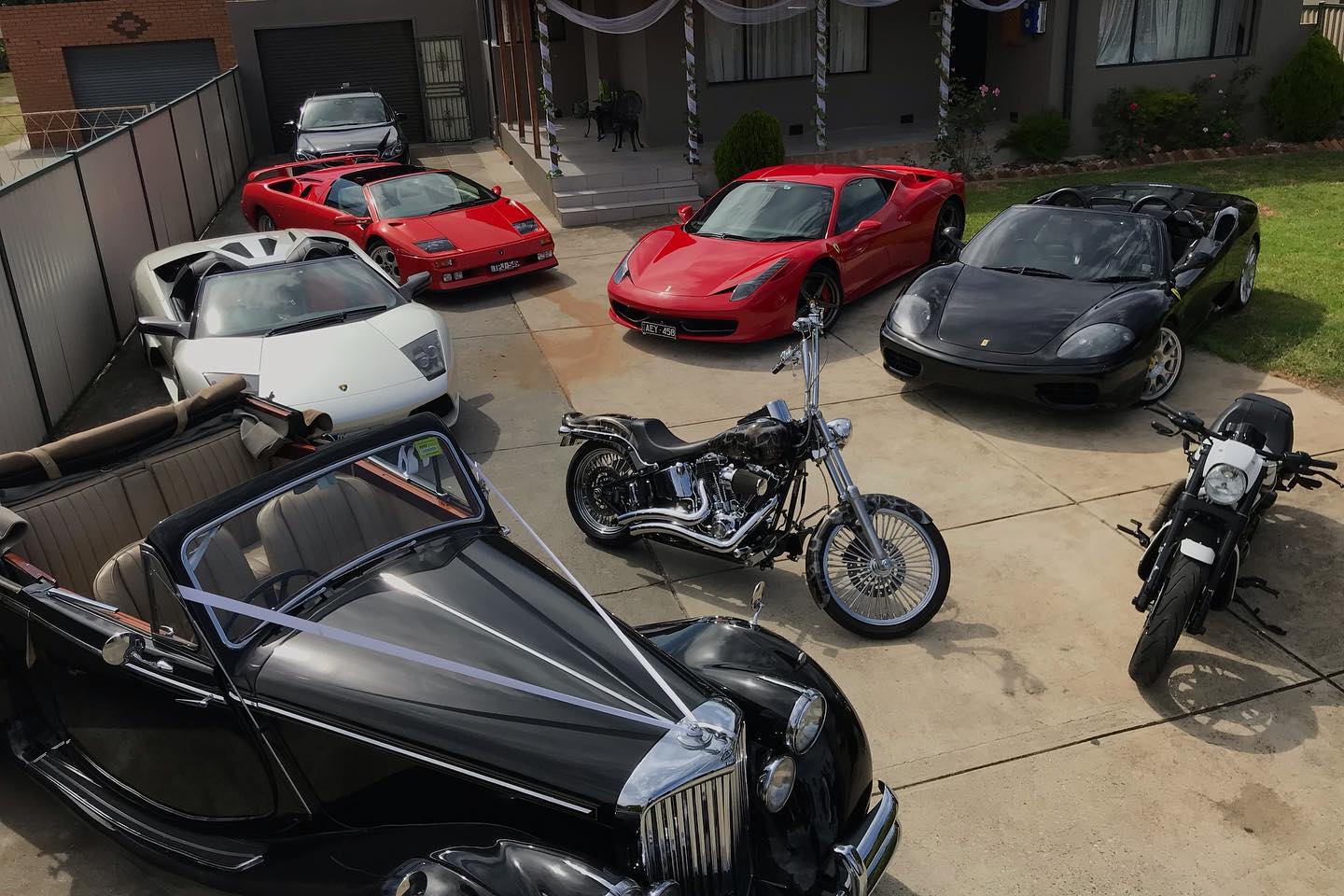 Black Harleys parked in a driveway with red and white sports cars and a vintage car