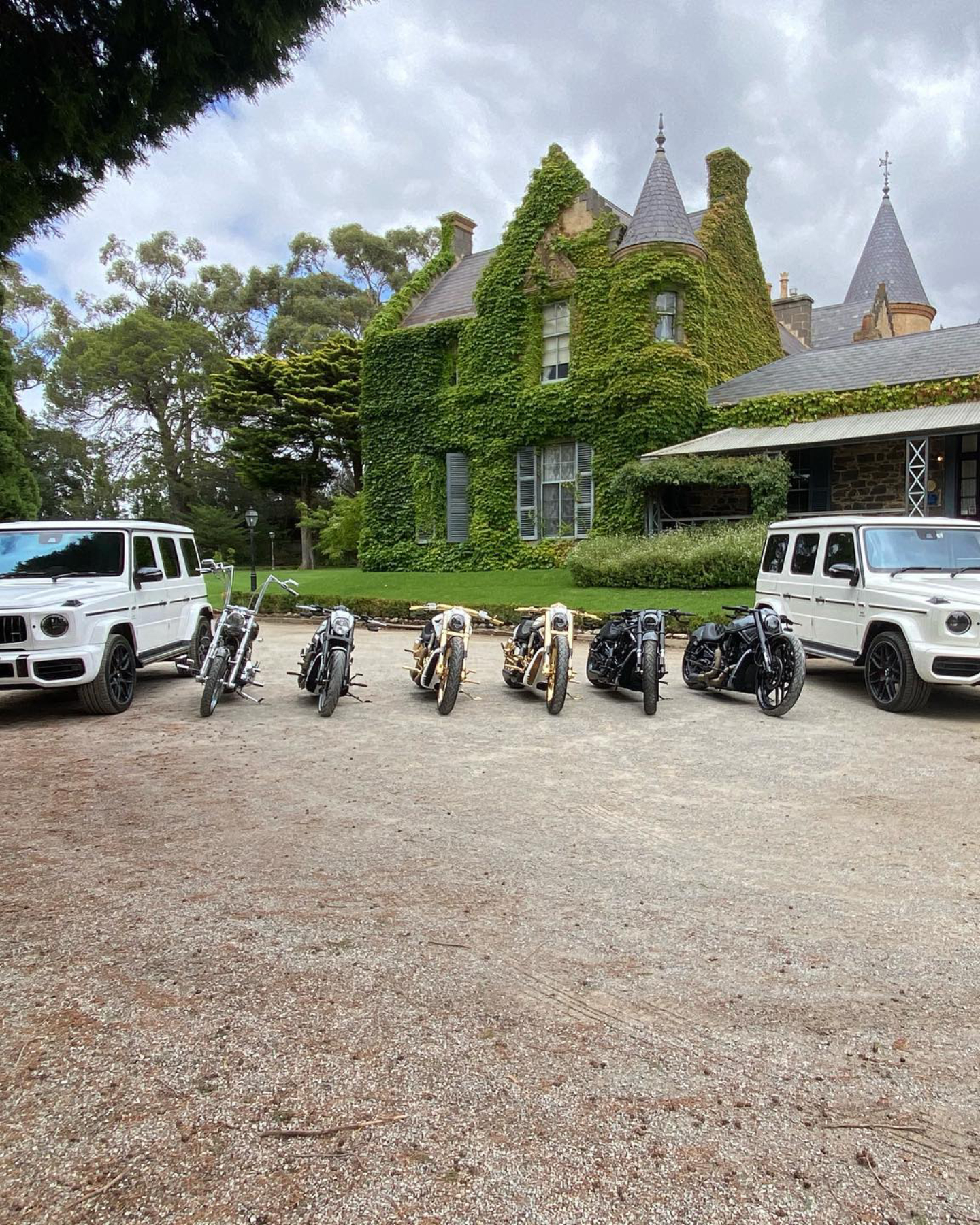 Six Harleys lined up between two white four-wheel drives outside an ivy-covered mansion