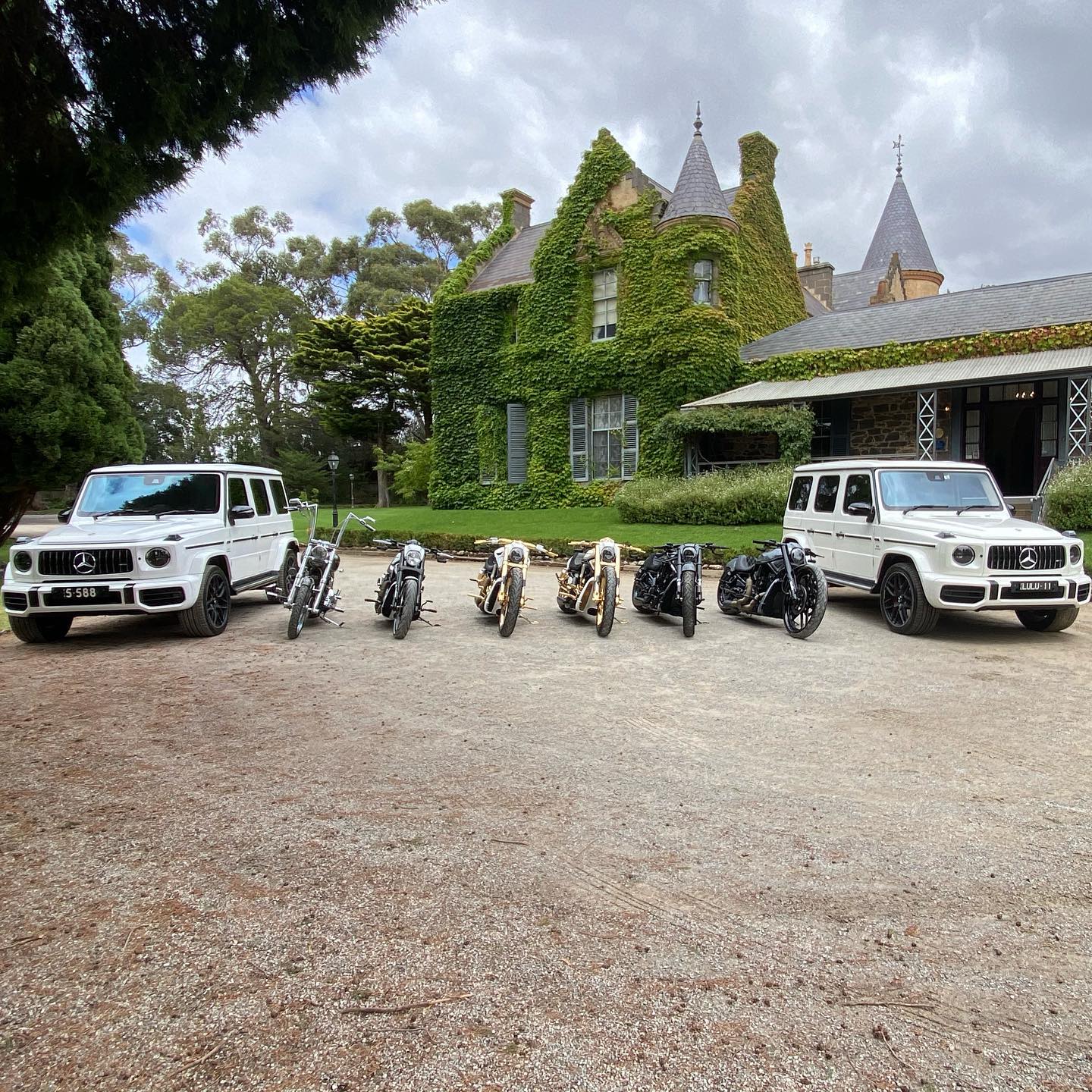 Harleys and two white four-wheel drives lined up on gravel outside an ivy-covered mansion