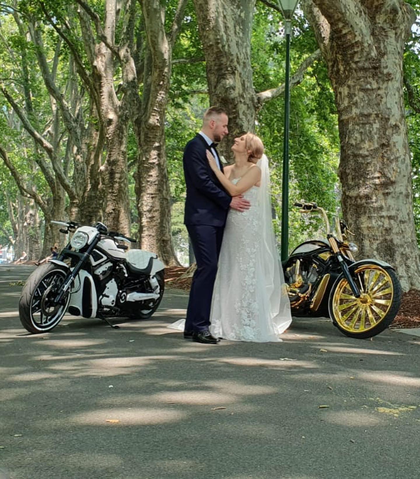 Bride and groom embracing between two Harleys on a tree-lined path