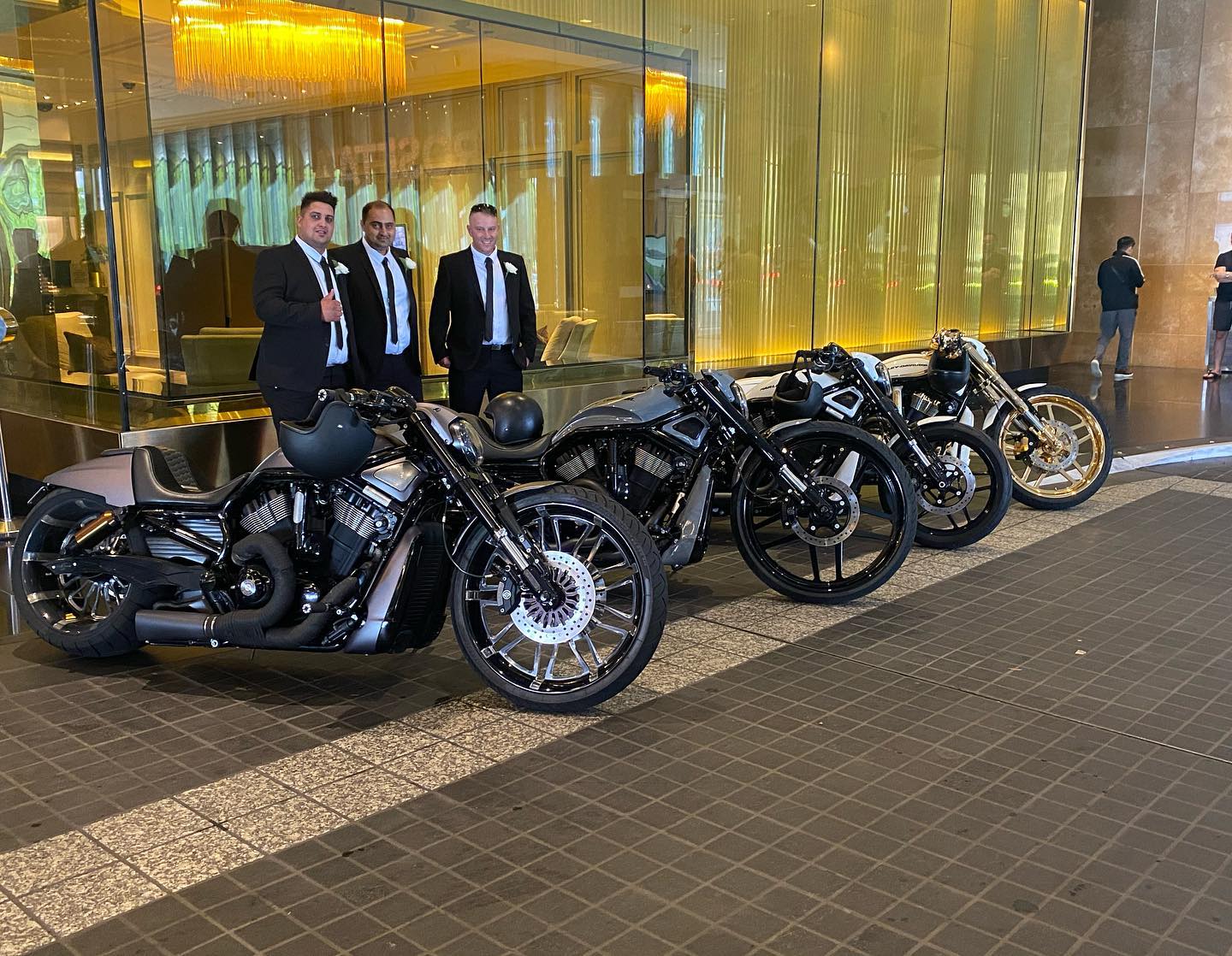 Three men in suits with a row of Harleys outside a hotel foyer