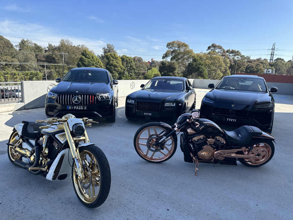 Gold-trimmed white Harley and copper-trimmed black Harley in front of three black luxury cars
