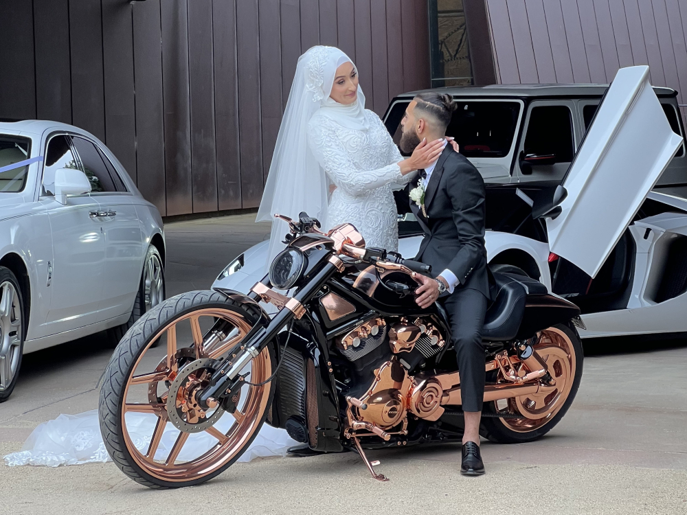 Bride and groom posing on a copper-trimmed black Harley beside luxury cars