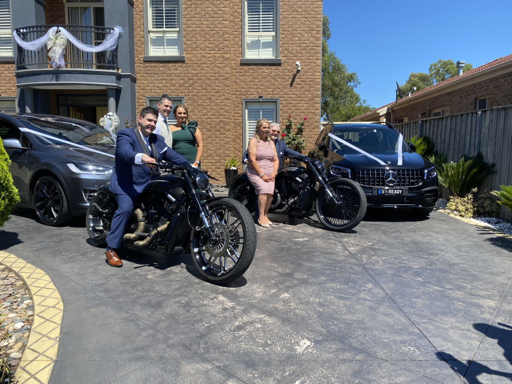 Couples posing with two black Harleys in a driveway with ribboned wedding cars