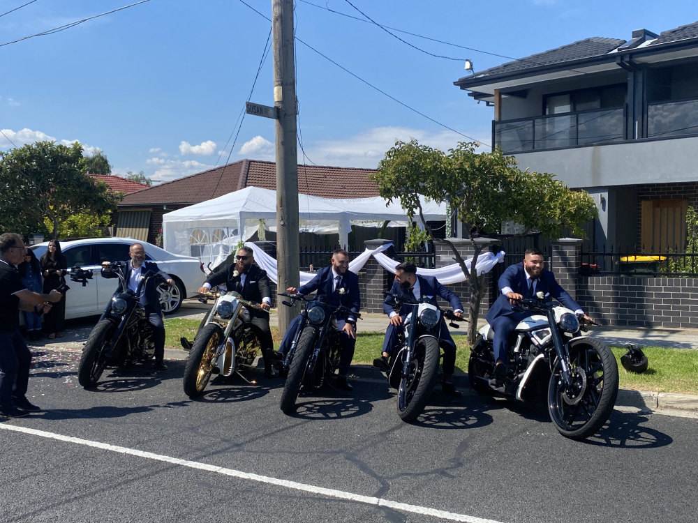 Groom and groomsmen in suits seated on a row of black Harleys in a street